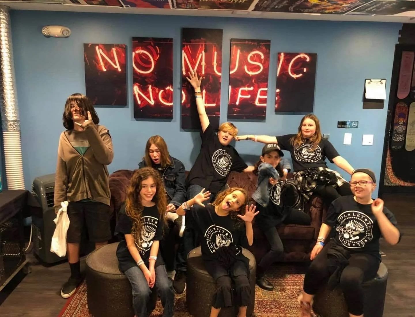 Let There Be Rock School students in the lounge beneath a NO MUSIC NO LIFE neon sign, most of them wearing the school's crest T-shirt