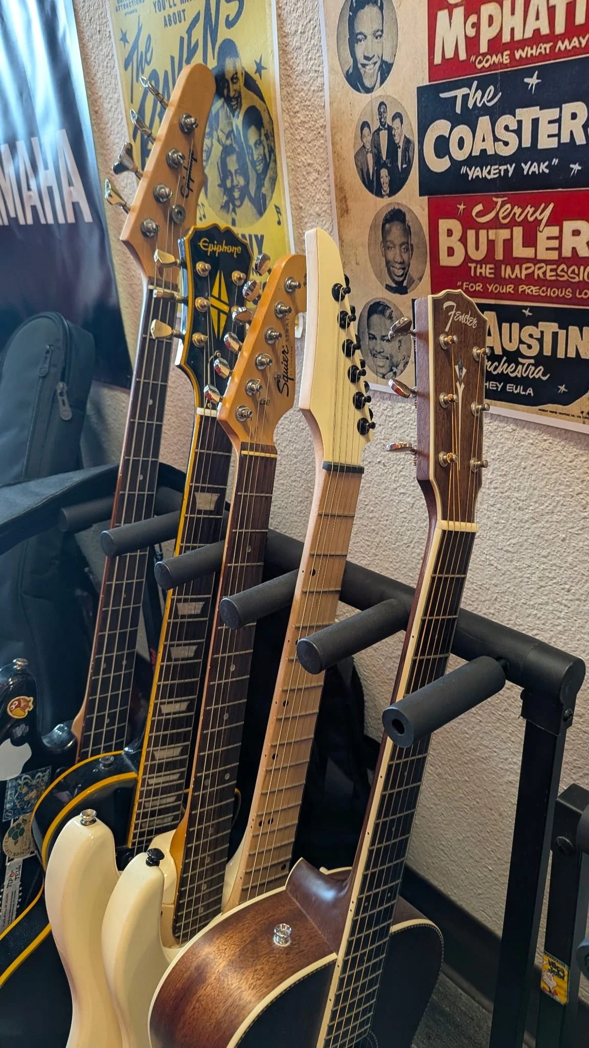 A rack of acoustic and electric guitars on the wall at Let There Be Rock School beside vintage concert posters