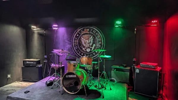 A Let There Be Rock School rehearsal room with a Yamaha drum kit, amplifiers and the school's crest painted on the wall, under magenta and green light