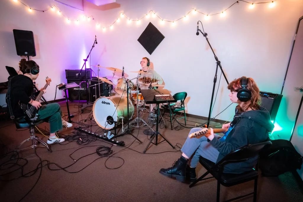 Three Let There Be Rock School students rehearsing together in a practice room strung with fairy lights, with a drum kit, bass and guitar