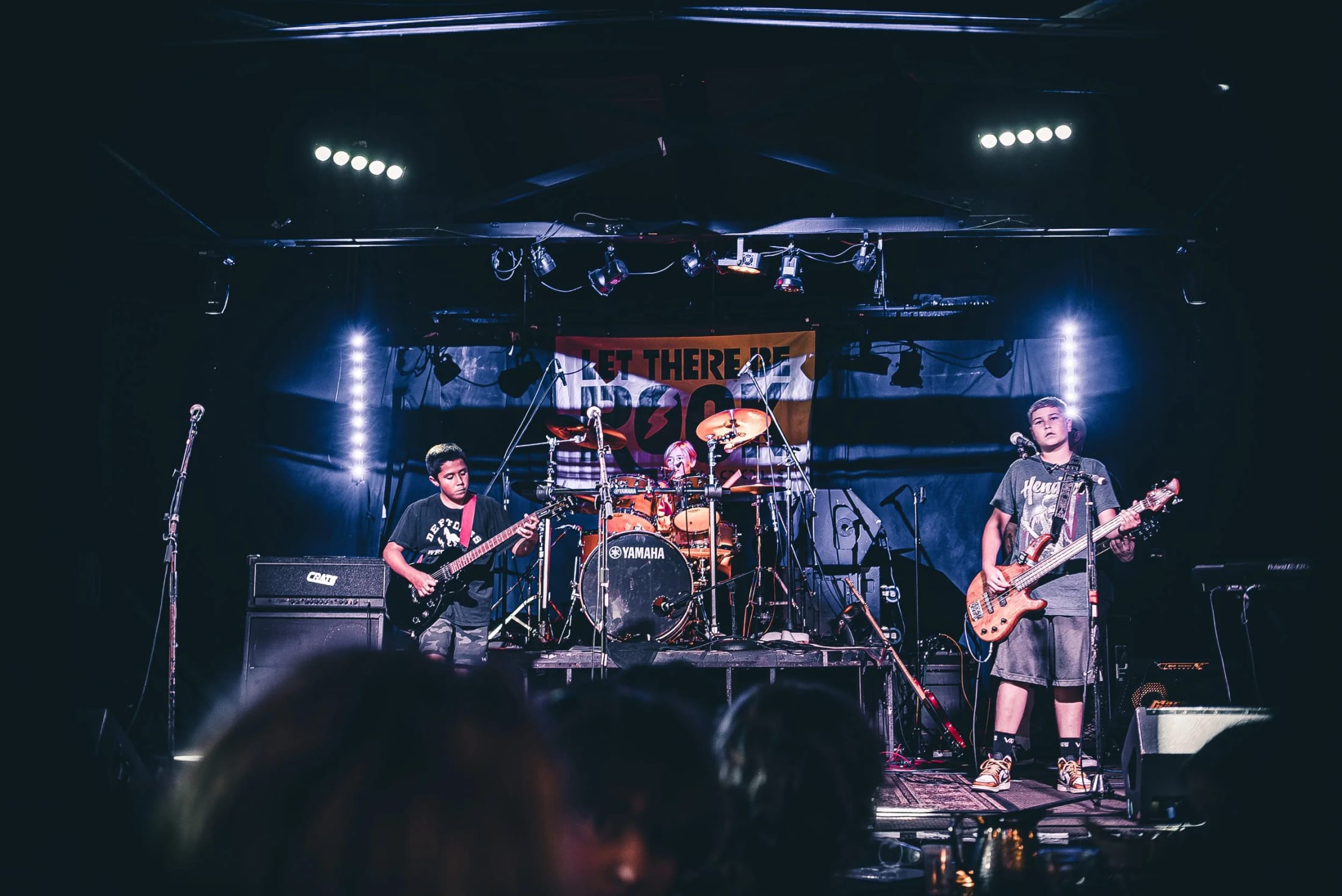 A student band playing on the Gaslamp stage in Long Beach beneath the orange LET THERE BE ROCK banner, audience heads silhouetted in the foreground
