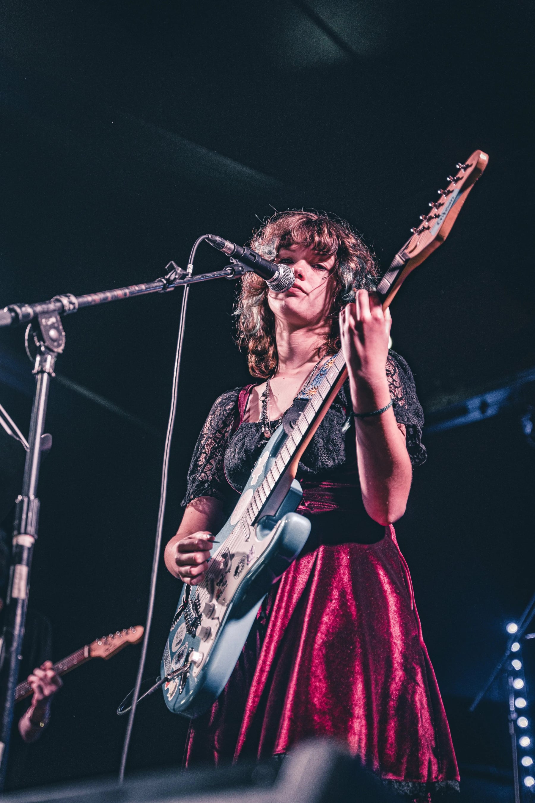 A Let There Be Rock School student singing into the microphone mid-song with an electric guitar, under stage lights