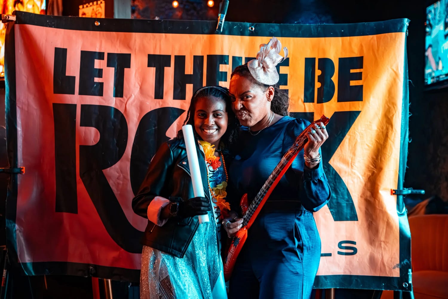 Two Let There Be Rock School students holding guitars in front of the orange LET THERE BE ROCK SCHOOLS stage banner