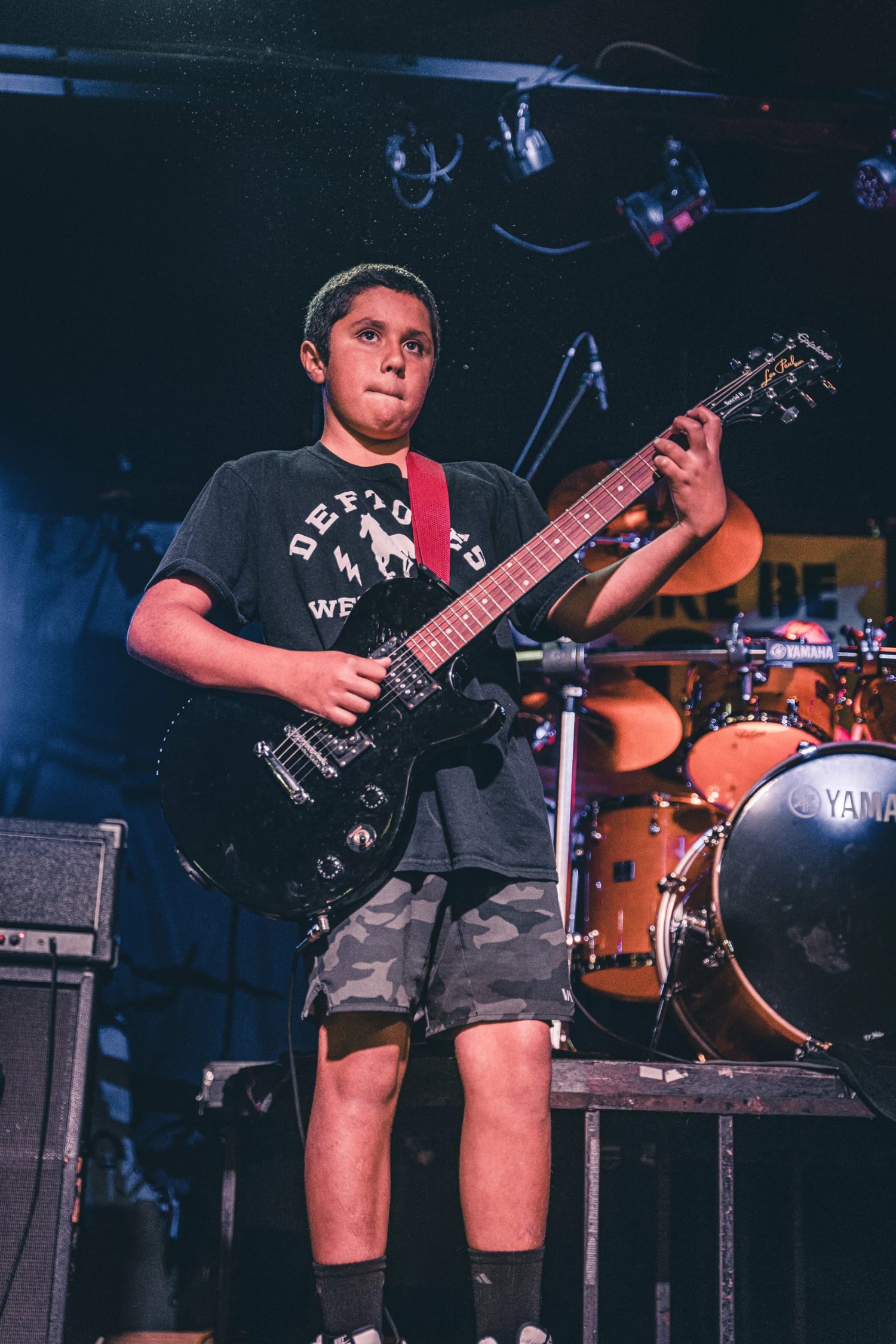A young student standing centre stage with a black electric guitar in front of the drum riser