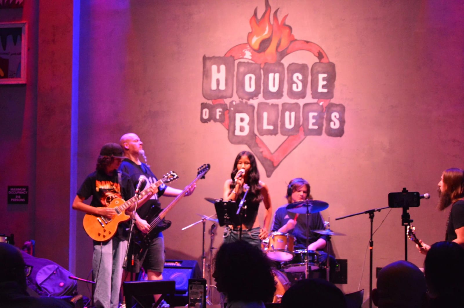 A student band performing on a club stage, singer at the mic, drummer and two guitarists behind